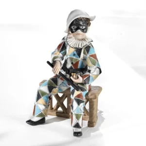 Statua in porcellana di Arlecchino in abito a toppe colorate, maschera nera e batòcio, seduto su una staccionata.