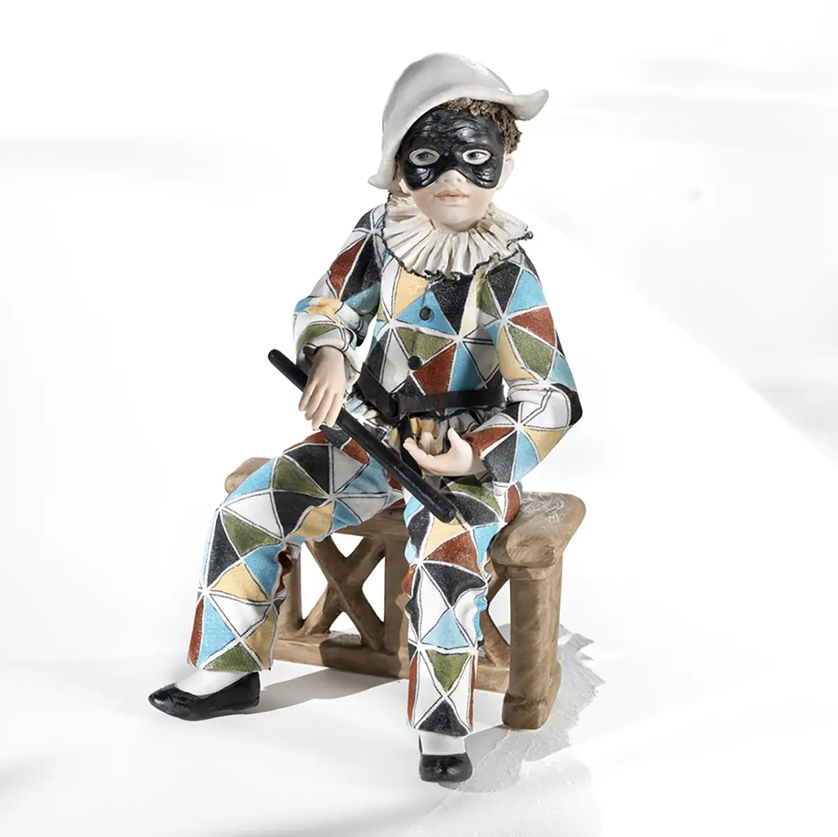 Statua in porcellana di Arlecchino in abito a toppe colorate, maschera nera e batòcio, seduto su una staccionata.