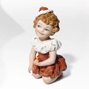 Statua in porcellana Chrissy in ginocchio con abito rosso, cappello natalizio e candelina dorata.