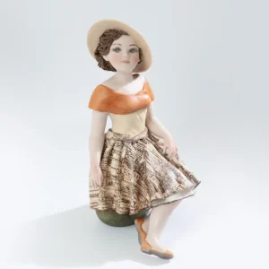 Statua in porcellana Deborah con abito burro, bavero arancione e ampio cappello, seduta su uno sgabello verde oliva.
