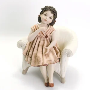 Figura in porcellana di ragazza seduta su poltrona in pelle bianca con abito rosa a fiori rossi e ballerine rosse.
