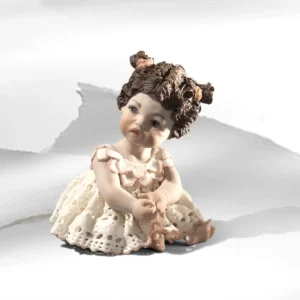 Statua in porcellana di bimba seduta a terra con abitino in pizzo dai bordi rosa e capelli legati in due codine alte.