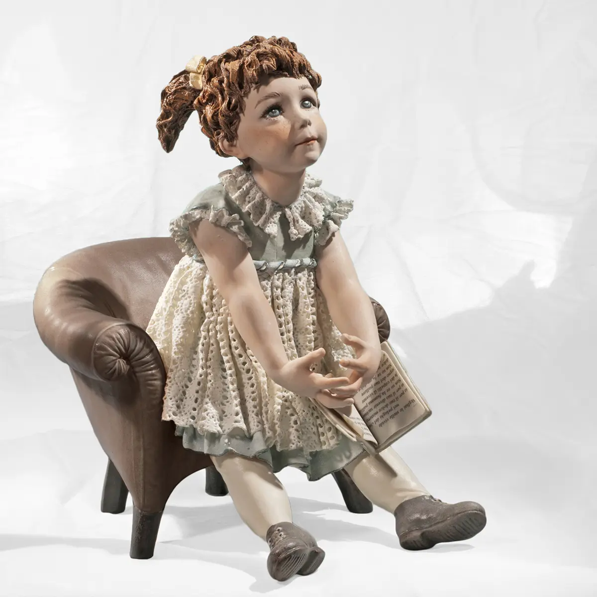 Statua in porcellana di bimba su poltrona pesca con abitino azzurro in pizzo e libro in grembo.