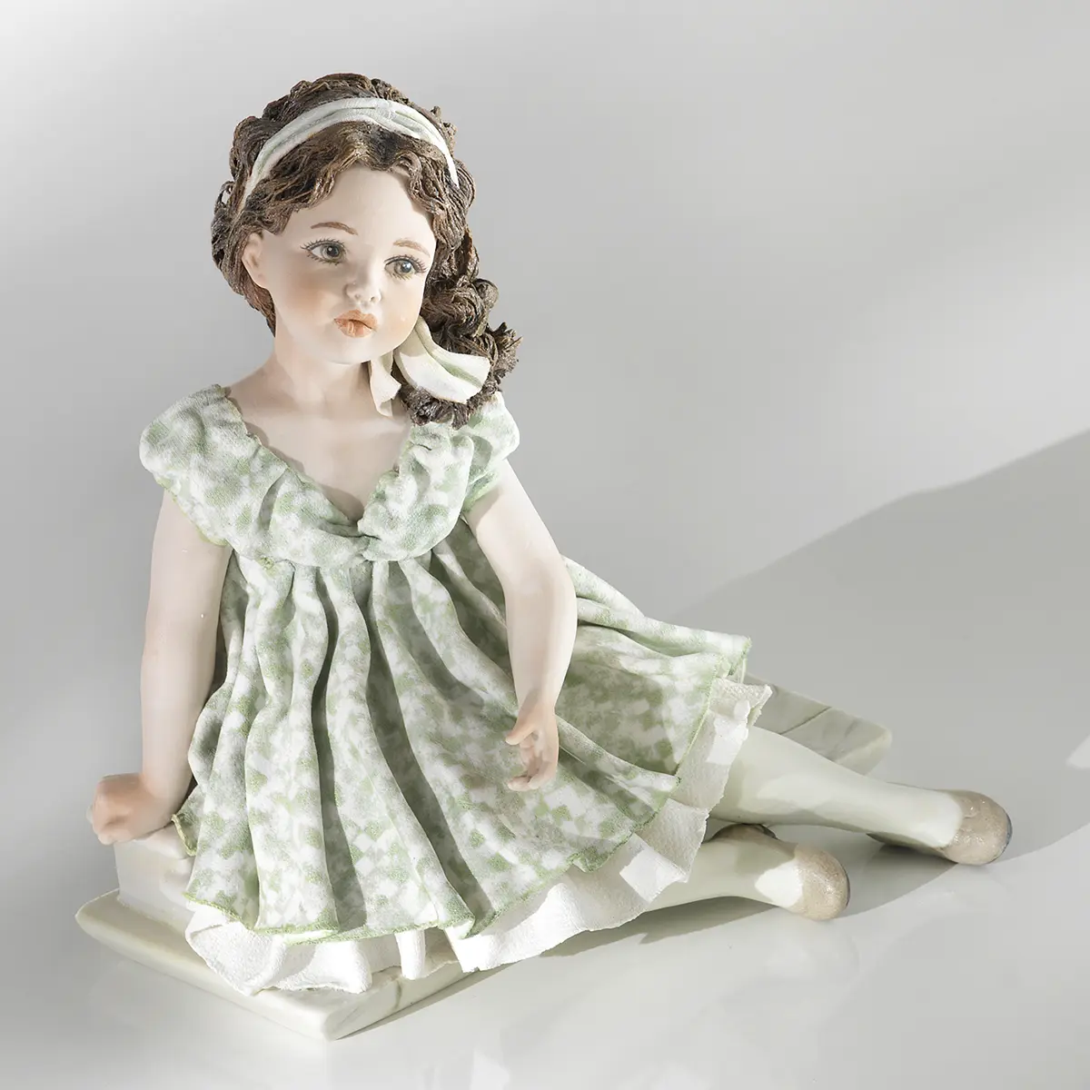 Statua in porcellana di ragazza in abito verde pastello seduta su scalino marmorizzato con fascia bianca nei capelli.