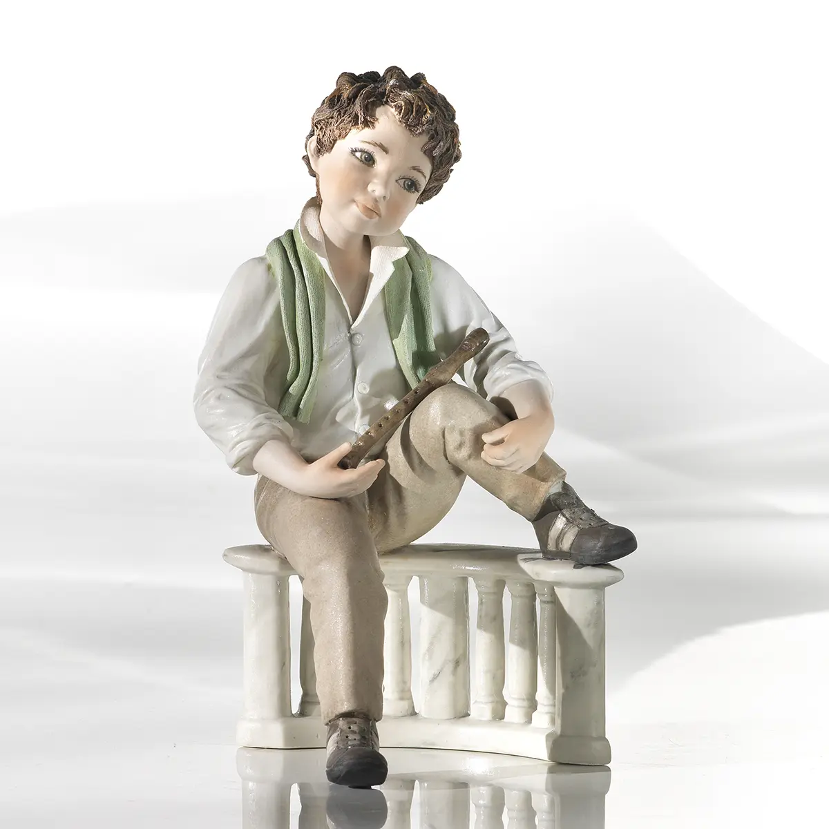 Statua in porcellana di ragazzo seduto su balaustra con camicia bianca, pantaloni marroni, sciarpa verde e flauto.