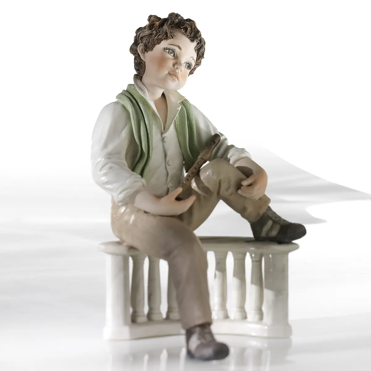 Statua in porcellana di ragazzo con flauto, sciarpa verde pastello e camicia bianca seduto su balaustra marmorizzata.