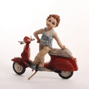Statua in porcellana di un ragazzino in piedi su una Vespa rossa con canottiera a righe bianche e azzurre e pantaloni beige.