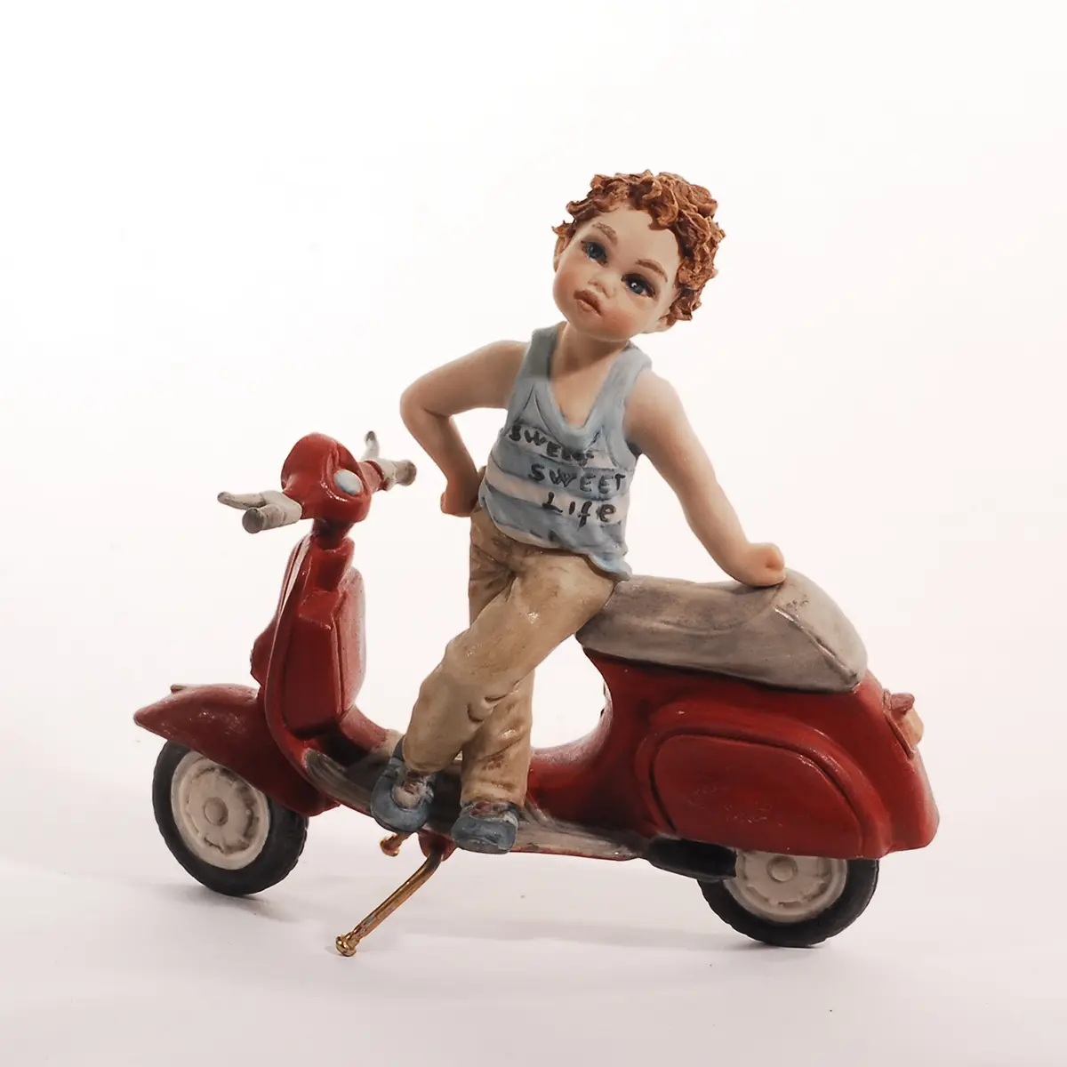 Statua in porcellana di un ragazzino in piedi su una Vespa rossa con canottiera a righe bianche e azzurre e pantaloni beige.