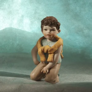 Statua in porcellana di Julien, bimbo in ginocchio con t-shirt azzurra, maglioncino giallo sulle spalle e pantaloni marroni.