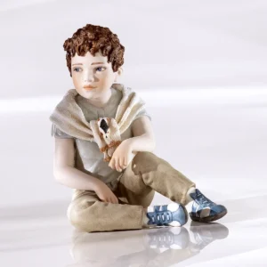 Statua in porcellana di ragazzo seduto a terra con pantaloni beige, maglietta azzurra e maglioncino a rombi sulle spalle.