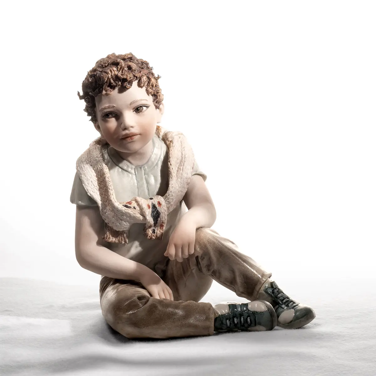 Statua in porcellana di ragazzo seduto con pantaloni beige, maglietta azzurra e maglioncino panna a rombi sulle spalle.
