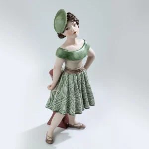 Statua in porcellana Maggie con gonna verde e bianca, bustino beige e cappellino fascinator, appoggiata a un tavolino.