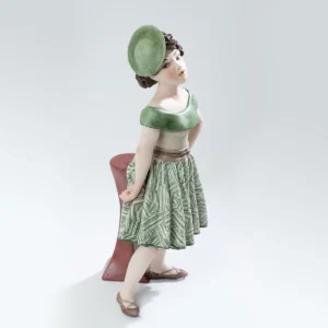 Statua in porcellana Maggie con gonna verde e bianca, bustino beige e cappello fascinator, appoggiata a un tavolino.