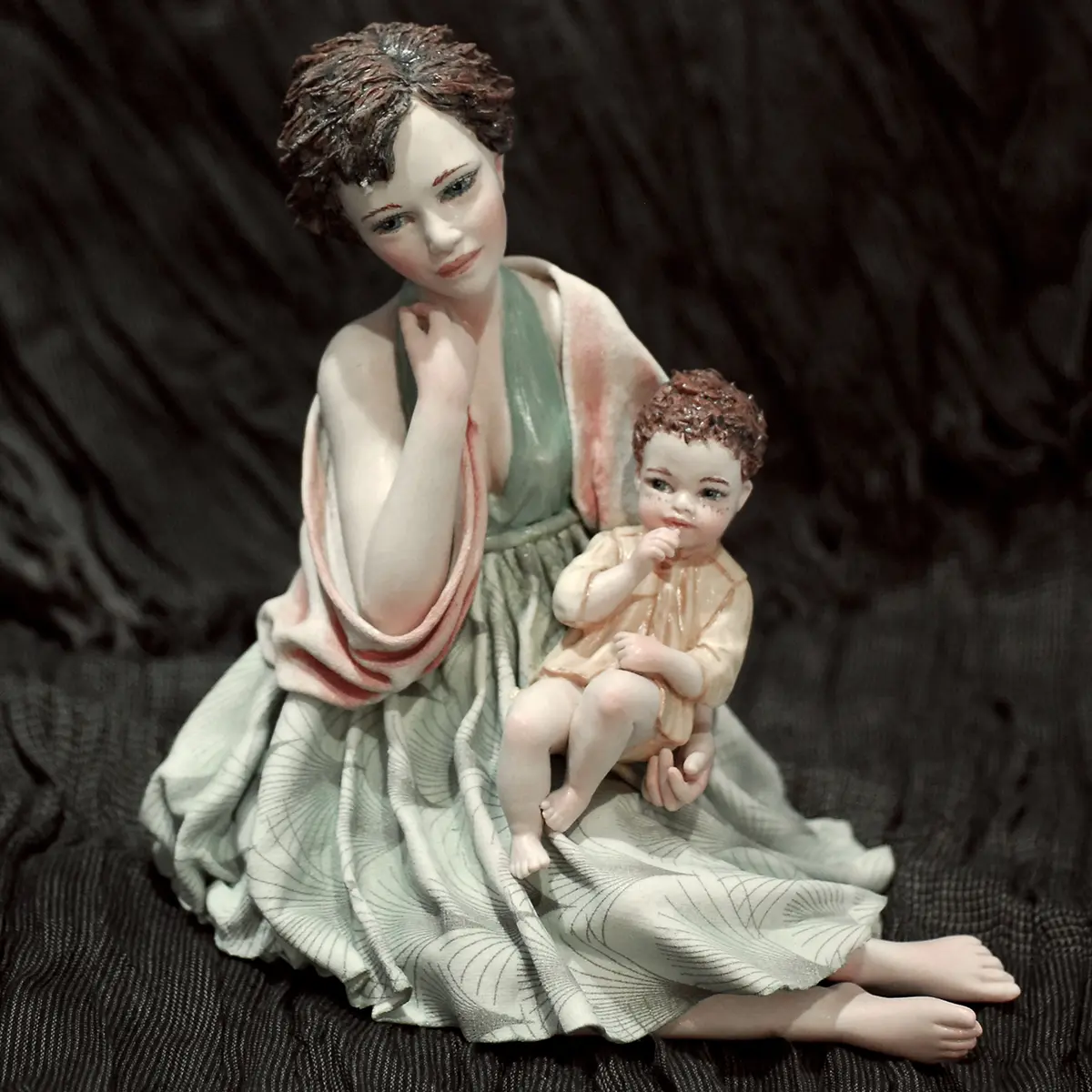 Statua in porcellana di mamma seduta con bimbo in braccio, in abito azzurro, scialle salmone e camiciola gialla.