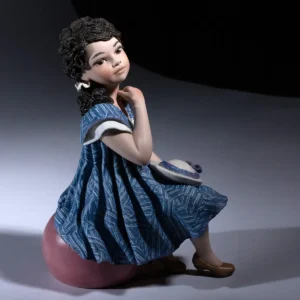 Statua in porcellana Nadia 2025 con abito indaco, seduta su pouf malva e cappellino bianco in grembo.