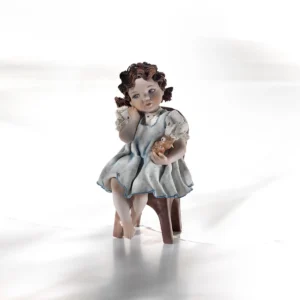 Statua in porcellana di bimba seduta su una seggiolina con abitino azzurro e orsetto giocattolo in braccio.