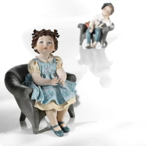 Statua in porcellana di bimba seduta su poltrona grigia con abitino azzurro, scarpine blu e codine con ponpon arancioni.