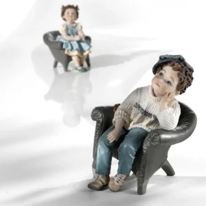 Statua in porcellana di bimbo seduto su poltrona grigia con jeans, maglioncino panna e berretto azzurro.