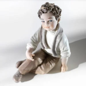 Statua in porcellana di bimbo seduto con maglioncino verde ghiaccio, pantaloni marroncini e sciarpa a quadretti.