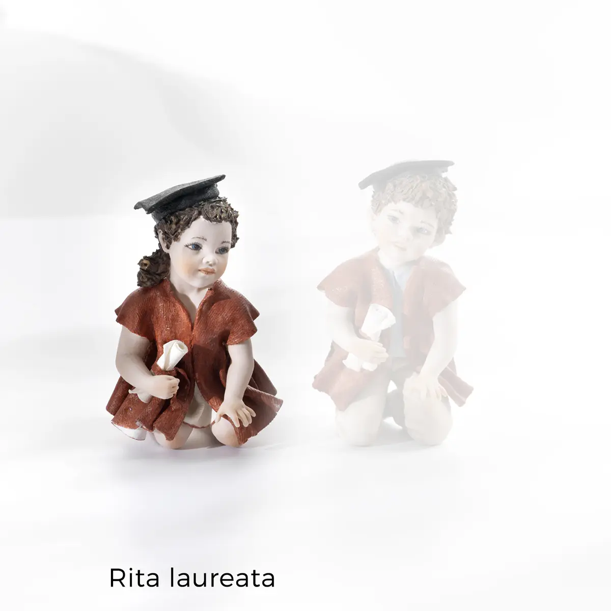 Statua in porcellana di Rita laureata con toga rossa, tocco nero e pergamena arrotolata.