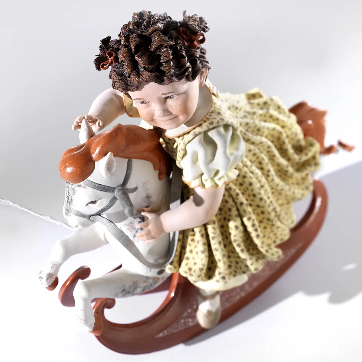 Statua in porcellana di bimba su cavallino a dondolo con abito giallo a fiorellini e cavallo con criniera rossa.