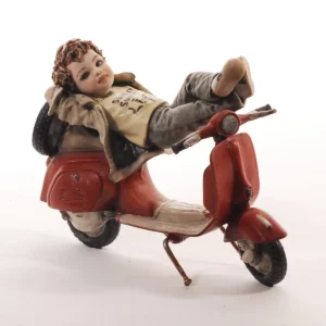 Statua in porcellana di ragazzino steso con i piedi sul volante di una Vespa rossa, con maglia gialla e giacca fango.